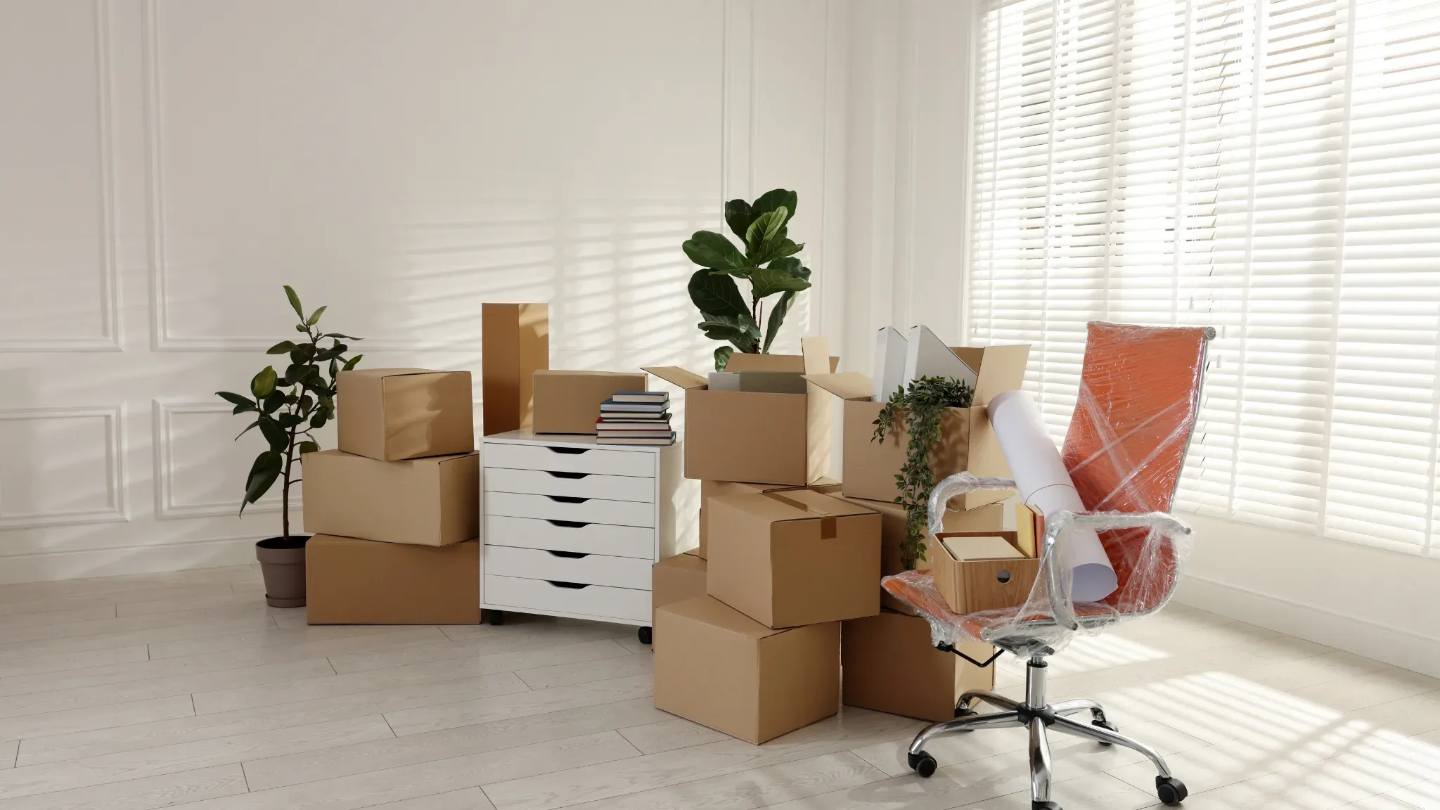 A bright, empty room mid-move is filled with packed cardboard boxes of varying sizes stacked around a white multi-drawer dres