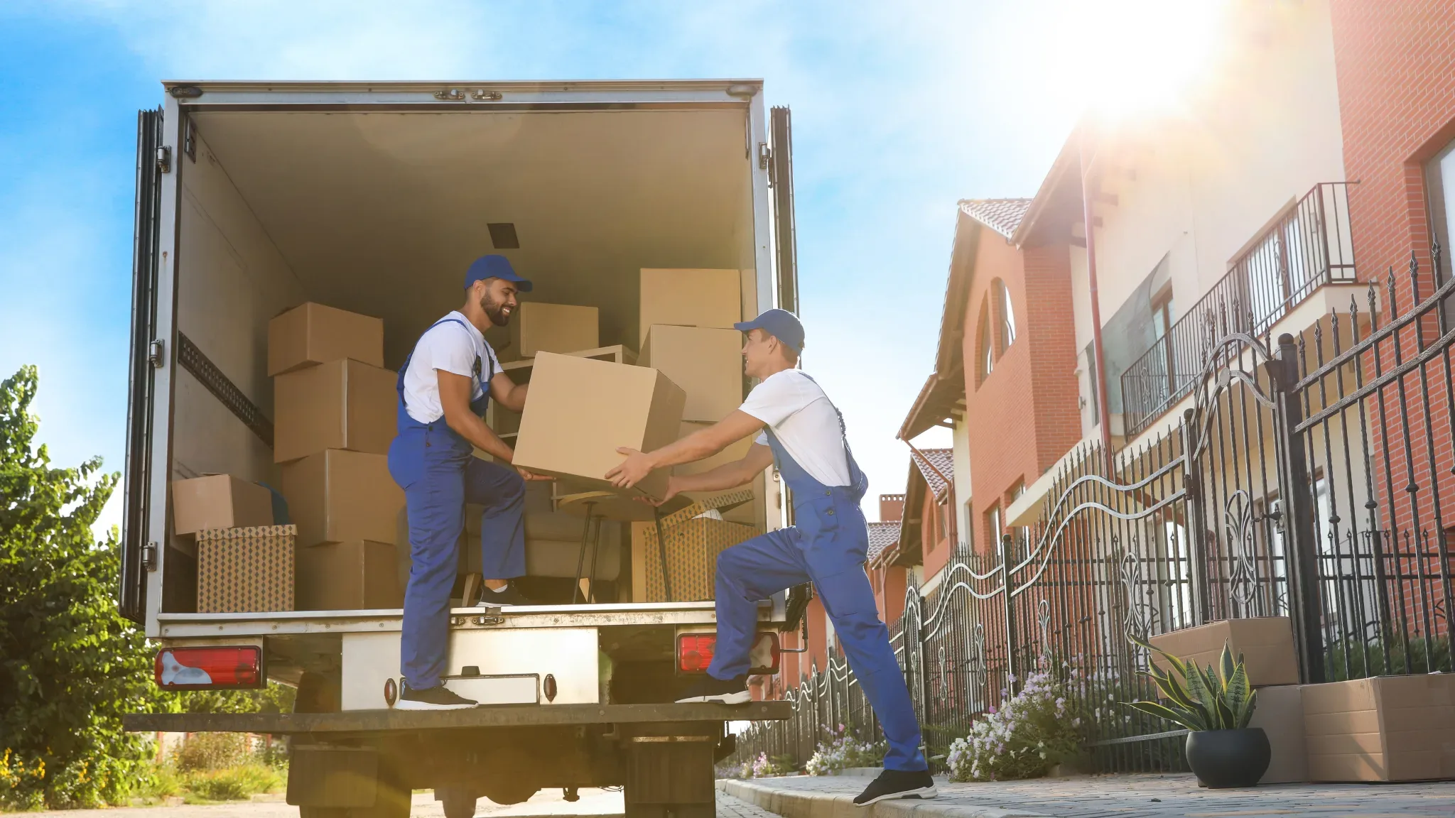 Two movers in matching blue coveralls and caps pass a cardboard box between them at the open back of a moving truck parked on