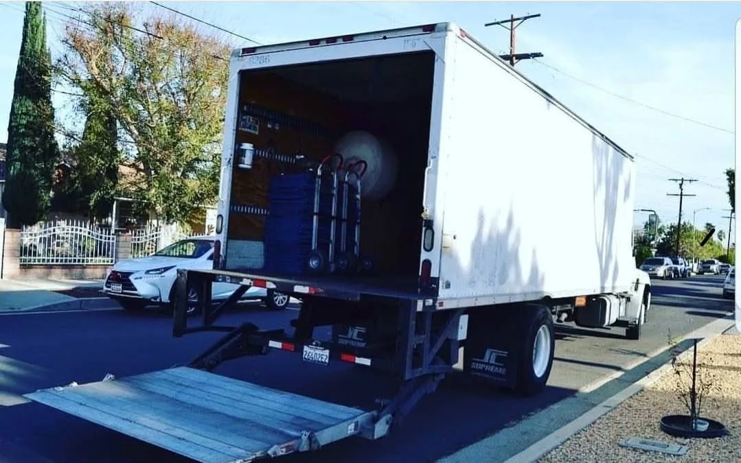 White moving truck with open cargo bay and lowered ramp parked on street with white car nearby.